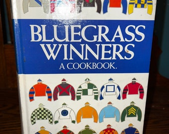 Könnte beinhalten: Ein weißes Kochbuch mit dem Titel "Bluegrass Winners: A Cookbook" mit einem blauen Streifen am unteren Rand. Der Umschlag zeigt bunte Jockey-Seiden in verschiedenen Mustern.
