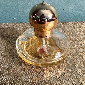 May include: A clear glass perfume bottle with a gold-colored, rounded top and a gold-colored base. The bottle is filled with a golden liquid and sits on a gold sequined surface. The bottle has a spray nozzle.