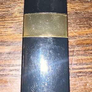 May include: A rectangular black lipstick case with a smooth, glossy finish and a wide, horizontal gold-colored metal band centered across the middle. The object has a flat top and bottom with slightly rounded corners and a reflective surface.