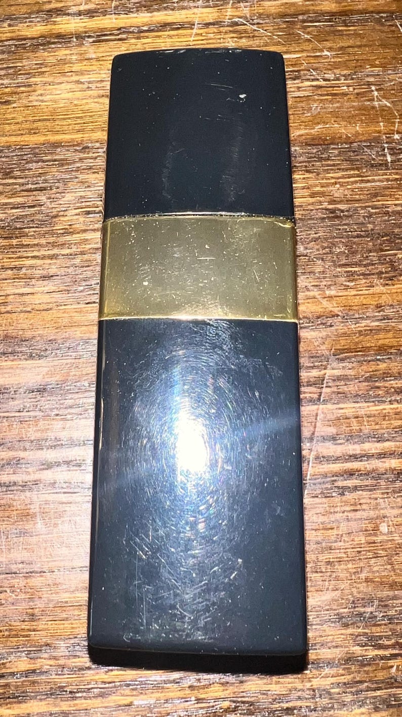 May include: A rectangular black lipstick case with a smooth, glossy finish and a wide, horizontal gold-colored metal band centered across the middle. The object has a flat top and bottom with slightly rounded corners and a reflective surface.
