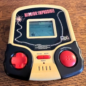 May include: A vintage handheld electronic game, featuring the title "MISSION: IMPOSSIBLE" in red text. The game has a black and tan color scheme, with a small screen displaying a pixelated action scene. It includes red control buttons and the "MGA" logo.