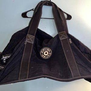 May include: Black nylon tote bag with two long shoulder straps and a white stitched trim. The bag features a circular logo patch with a star and the brand name. The bag is hanging on a black hanger.