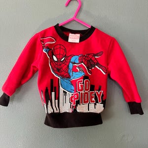K&ouml;nnte beinhalten: Ein rotes Langarm-Sweatshirt mit einer Grafik von Spider-Man in dynamischer Pose &uuml;ber einer schwarz-wei&szlig;en Stadtsilhouette. Der Text GO SPIDEY ist in Rot und Wei&szlig; gedruckt. Das Kleidungsst&uuml;ck hat einen schwarzen Rundhalsausschnitt und schwarze Rippb&uuml;ndchen.
