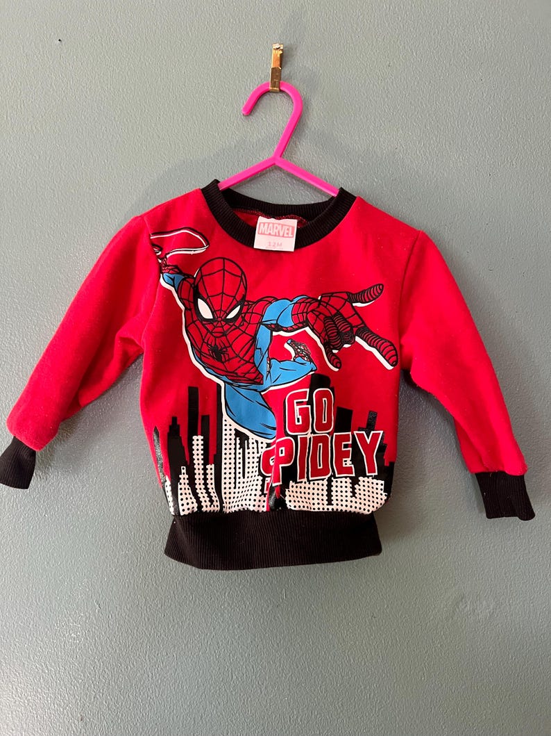 K&ouml;nnte beinhalten: Ein rotes Langarm-Sweatshirt mit einer Grafik von Spider-Man in dynamischer Pose &uuml;ber einer schwarz-wei&szlig;en Stadtsilhouette. Der Text GO SPIDEY ist in Rot und Wei&szlig; gedruckt. Das Kleidungsst&uuml;ck hat einen schwarzen Rundhalsausschnitt und schwarze Rippb&uuml;ndchen.