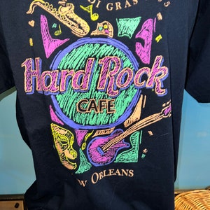 May include: Black t-shirt with a Hard Rock Cafe design for Mardi Gras 1995 in New Orleans. The graphic includes musical instruments and masks, with the Hard Rock Cafe logo in the centre, in a vibrant colour scheme.