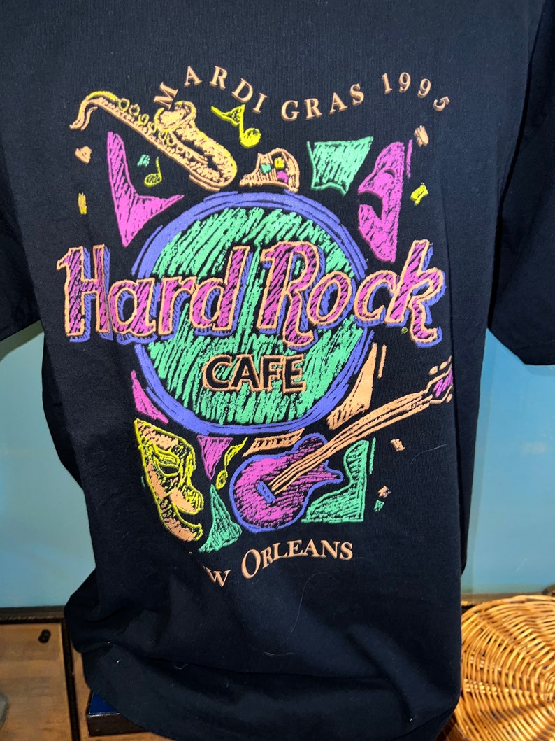 May include: Black t-shirt with a Hard Rock Cafe design for Mardi Gras 1995 in New Orleans. The graphic includes musical instruments and masks, with the Hard Rock Cafe logo in the centre, in a vibrant colour scheme.