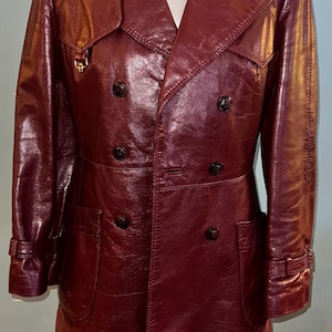 May include: A burgundy leather trench coat with a double-breasted design. The coat features a notched collar, two front pockets, and black buttons. The sleeves have adjustable cuffs with buckles. The coat is displayed on a mannequin.