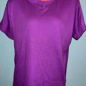 May include: A short-sleeved, purple ribbed knit shirt with a round neckline and a single button closure. The shirt has a simple, classic design and is made of a soft, textured fabric. The color is a vibrant, solid purple.