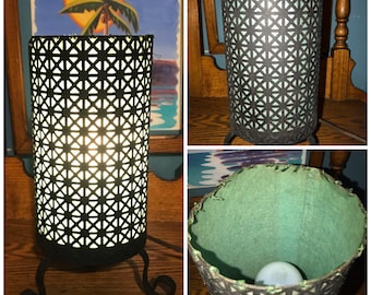 May include: A black metal table lamp with a green fabric shade. The shade has a geometric pattern and is lit by a light bulb. The lamp has a three-legged base.