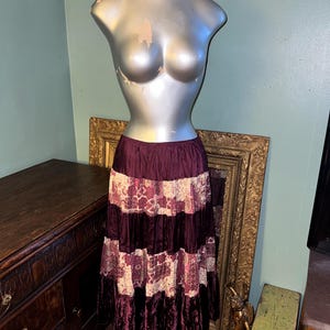 May include: A tiered, floor-length skirt in shades of burgundy and cream, displayed on a mannequin. The skirt features a velvet texture with patterned panels and a ruffled hem, showcasing a bohemian style.