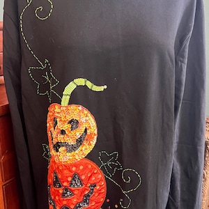 Puede incluir: Una camiseta negra de manga larga con dos calabazas de Halloween apiladas, decoradas con lentejuelas naranjas y rojas. El diseño incluye enredaderas y hojas verdes bordadas, con pequeñas cuentas de colores esparcidas cerca del dobladillo.