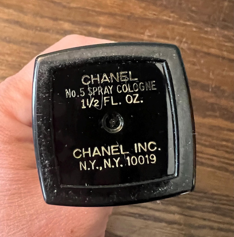 May include: A black rectangular base of a Chanel No.5 spray cologne bottle with gold lettering that reads CHANEL, No.5 SPRAY COLOGNE, 1 1/2 FL. OZ., CHANEL INC., N.Y., N.Y. 10019, featuring a central circular spray nozzle mechanism on the surface.