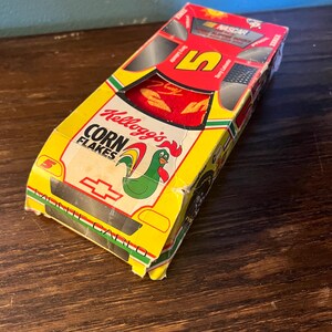 May include: A vintage Kellogg's Corn Flakes box designed as a race car. The box is yellow, red, and green, featuring the number 5 and the Kellogg's rooster logo. The box is labeled "Monte Carlo" and "NASCAR".