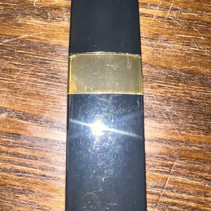 May include: A rectangular lipstick case with a smooth black finish and a central horizontal gold-toned band. The casing features a sleek, minimalist design with a reflective surface that shows light glints and minor surface scratches across the body.