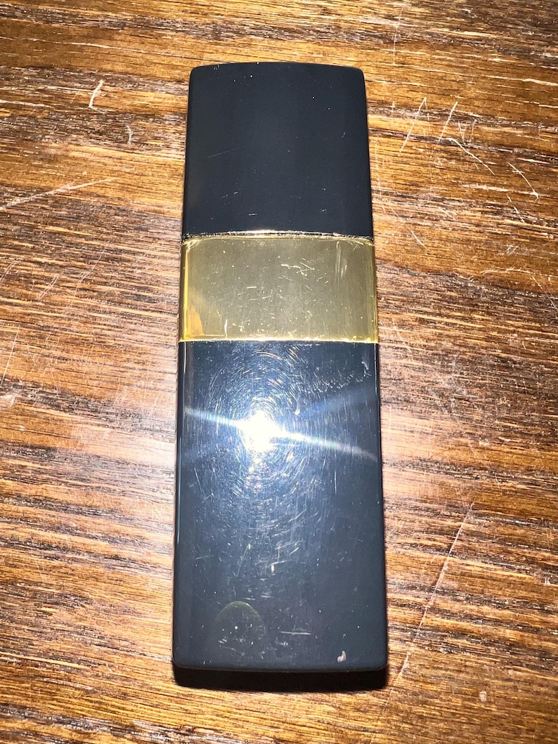 May include: A rectangular lipstick case with a smooth black finish and a central horizontal gold-toned band. The casing features a sleek, minimalist design with a reflective surface that shows light glints and minor surface scratches across the body.