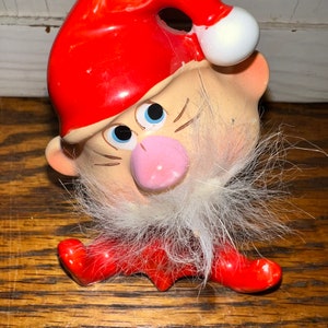 May include: A ceramic Christmas gnome figurine with a red hat, white beard, and a pink nose. The gnome is sitting with its legs crossed and has a mischievous expression on its face.