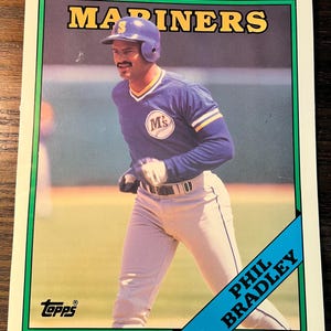 May include: A vintage baseball card featuring Phil Bradley of the Seattle Mariners. The card has a green border with the team name "MARINERS" in yellow. Bradley is in a blue uniform with the team logo, and the card includes the Topps logo.