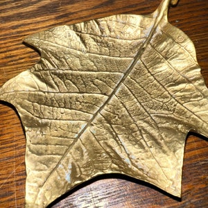 May include: A gold-toned brass leaf-shaped dish with a textured surface. The dish has a raised handle on one side.