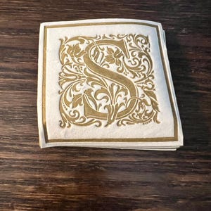 May include: A stack of square, off-white paper coasters with a gold-colored, ornate letter "S" design. The letter is surrounded by decorative floral and leaf patterns, with a gold border. The coasters are stacked on a dark brown wooden surface.
