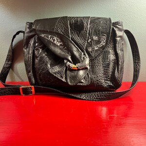 May include: A black leather shoulder bag with a unique embossed design and a gold buckle. The bag has a long strap and a flap closure.