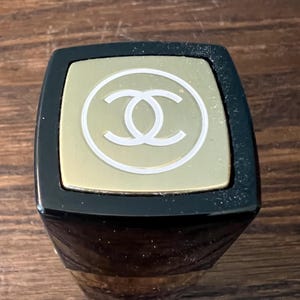 May include: A square lipstick tube cap featuring a black border and a gold-toned top panel. The center of the gold panel displays a white circular outline containing the interlocking double C logo. The design is viewed from a top-down perspective.