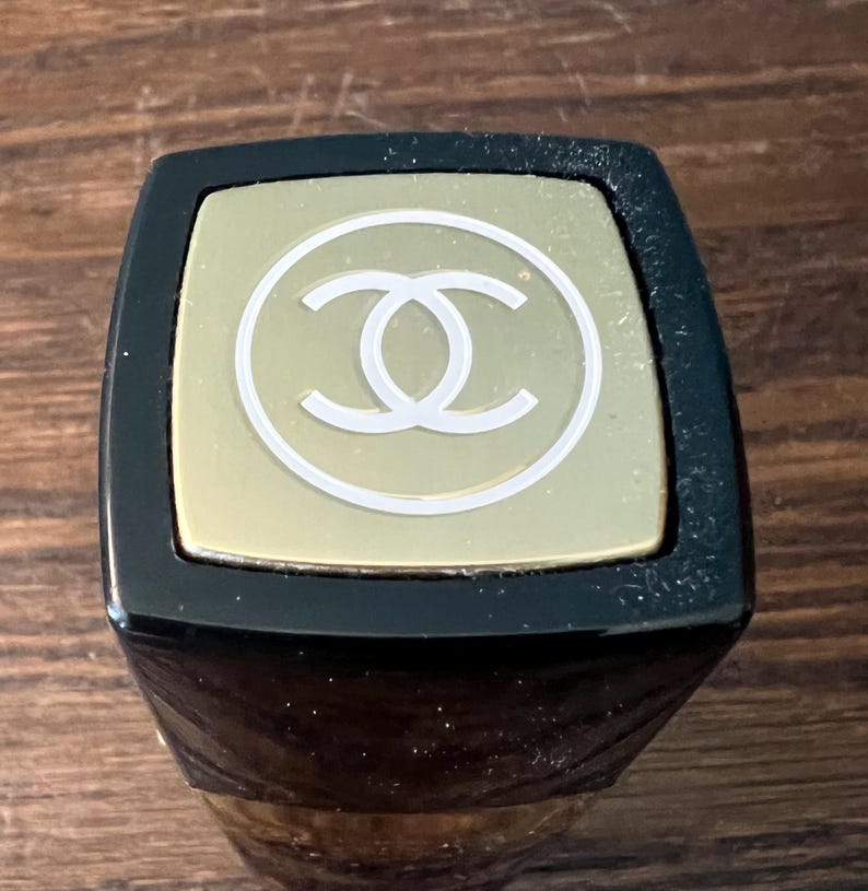 May include: A square lipstick tube cap featuring a black border and a gold-toned top panel. The center of the gold panel displays a white circular outline containing the interlocking double C logo. The design is viewed from a top-down perspective.