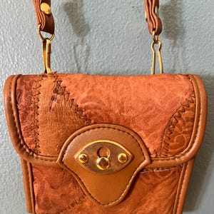 May include: A small brown leather crossbody bag featuring a textured, embossed pattern on the front flap. The bag includes a gold-toned metal clasp closure, smooth leather trim, and a matching brown leather strap attached with gold-toned metal hardware.
