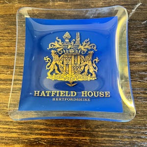 May include: A square glass ashtray with a blue center and clear edges. The blue center features a gold crest with the words "HATFIELD HOUSE HERTFORDSHIRE". The ashtray has a slightly curved shape.