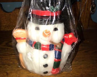 May include: A white snowman candle with a black top hat, red scarf, and a green and blue patterned scarf. The candle is wrapped in clear plastic with a red bow.
