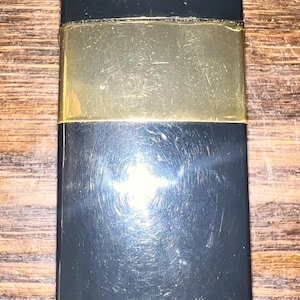 May include: A rectangular lipstick case with a glossy black finish and a wide, horizontal gold-toned metal band encircling the center. The surface of the black casing shows fine, visible scratches across the material, reflecting light.