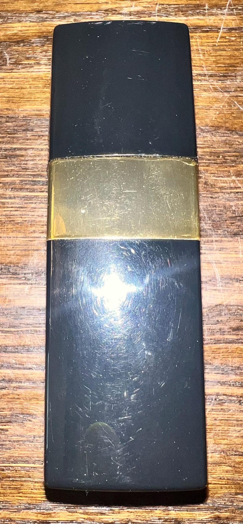 May include: A rectangular lipstick case with a glossy black finish and a wide, horizontal gold-toned metal band encircling the center. The surface of the black casing shows fine, visible scratches across the material, reflecting light.