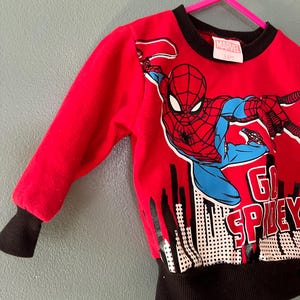 K&ouml;nnte beinhalten: Ein rotes Langarm-Sweatshirt mit einer Grafik der Marvel-Figur Spider-Man in dynamischer Pose &uuml;ber einer schwarz-wei&szlig;en Skyline. Das Oberteil zeigt den Schriftzug GO SPIDEY sowie einen schwarzen gerippten Rundhalsausschnitt und schwarze &Auml;rmelb&uuml;ndchen.