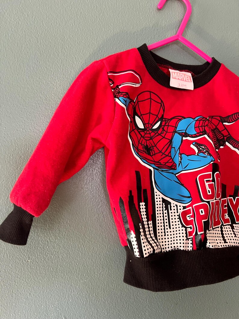 K&ouml;nnte beinhalten: Ein rotes Langarm-Sweatshirt mit einer Grafik der Marvel-Figur Spider-Man in dynamischer Pose &uuml;ber einer schwarz-wei&szlig;en Skyline. Das Oberteil zeigt den Schriftzug GO SPIDEY sowie einen schwarzen gerippten Rundhalsausschnitt und schwarze &Auml;rmelb&uuml;ndchen.
