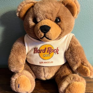 May include: A brown teddy bear wearing a white tank top with the Hard Rock Cafe logo and "LOS ANGELES" printed on it. The bear has a black nose and eyes, and is sitting on a wooden surface.