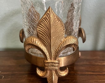 May include: A clear glass votive candle holder with a crackled texture, held in a gold-colored metal frame. The frame features a fleur-de-lis design. The candle holder sits on a dark wooden surface.