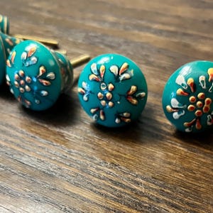 May include: A collection of teal-colored cabinet knobs with floral designs in white, orange, and gold. The knobs have a round shape and are arranged on a wooden surface, showcasing their decorative appeal.
