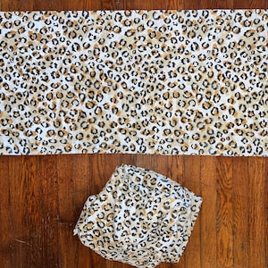 May include: A white fabric with a brown and black leopard print pattern. The fabric is folded in half and laid flat on a wooden surface.