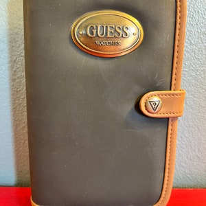 May include: A dark gray Guess Watches case with a brown leather trim and a gold-colored metal Guess Watches emblem. The case has a snap closure and is rectangular in shape.