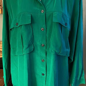 May include: A long-sleeved button-down shirt in a vibrant emerald green color. The shirt features a classic collar, two large chest pockets with flaps, and a central vertical row of dark buttons running down the front placket of the garment.