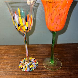 May include: Two colorful glass wine glasses. The glass on the left has a clear base with a colorful confetti design. The glass on the right has a green stem and a bright orange bowl with a speckled design.