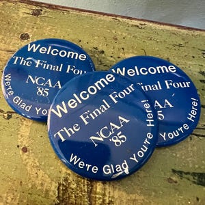 May include: Three blue round buttons with white text. The buttons say "Welcome The Final Four NCAA '85 We're Glad You're Here!" The buttons are on a distressed green surface.