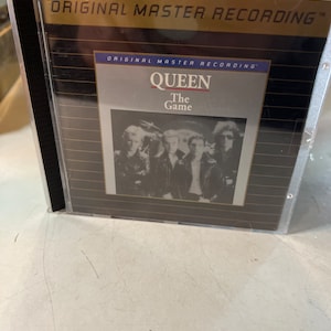 May include: A CD case with a gold background and black text that reads "Original Master Recording". The front of the case features a black and white photo of the band Queen with the text "Queen The Game" in white.