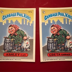 May include: Two Garbage Pail Kids trading cards featuring cartoon characters with the text "Garbage Pail Kids" and "141a" and "141b". The first card features a character named "Ashley Can" holding a garbage bag with the text "I ♥ NY". The second card features a character named "Greta Garbage" holding a garbage bag with the text "I ♥ NY".