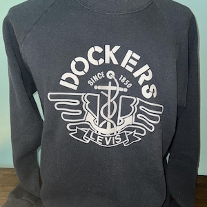May include: A dark gray long-sleeve sweatshirt featuring a white graphic print on the chest. The design includes the text DOCKERS, SINCE 1850, and LEVIS, surrounding an anchor and wing emblem. The garment has a crew neckline and raglan sleeves.