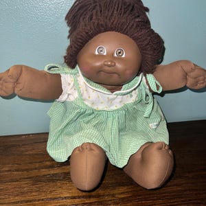 May include: A vintage Cabbage Patch Kid doll with dark skin, brown yarn hair, and brown eyes. The doll is wearing a green and white gingham dress with a white floral print collar. The doll is sitting with its arms and legs outstretched.