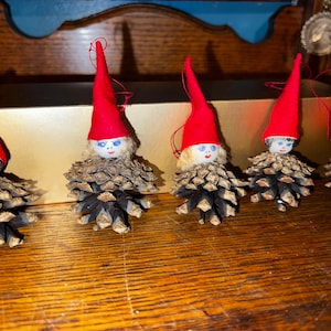 May include: Five small pine cone ornaments with red felt hats and painted faces. The ornaments have blue eyes and black hair.