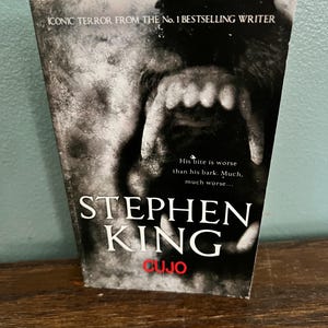 May include: A paperback book titled "CUJO" by Stephen King. The cover features a menacing dog's face with exposed teeth. The text reads "ICONIC TERROR FROM THE No. 1 BESTSELLING WRITER" and "His bite is worse than his bark. Much, much worse..."