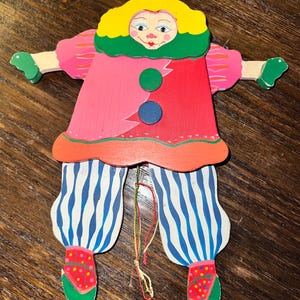 May include: A colorful wooden clown toy with a red hat, yellow hair, and a painted face. The clown wears a pink and red outfit with blue and white striped pants. The arms and legs are jointed, and the toy has a string for movement.