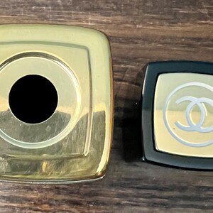 May include: A large square gold-toned cap with a central circular opening next to a smaller square black cap featuring a gold-toned top panel with a white interlocking CC logo emblem centered on the surface of the smaller cap.