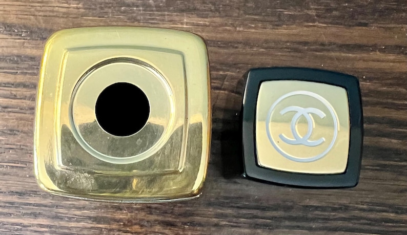 May include: A large square gold-toned cap with a central circular opening next to a smaller square black cap featuring a gold-toned top panel with a white interlocking CC logo emblem centered on the surface of the smaller cap.